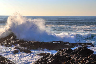 Waves splashing on rocks at shore against sky