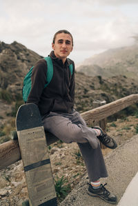 Portrait of young man sitting on railing by skateboard