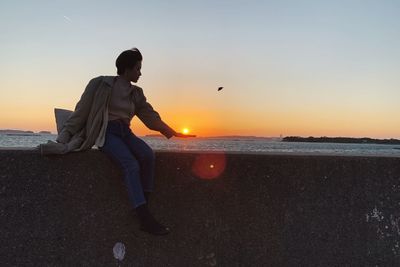 Full length of woman sitting on retaining wall against sea and sky during sunset