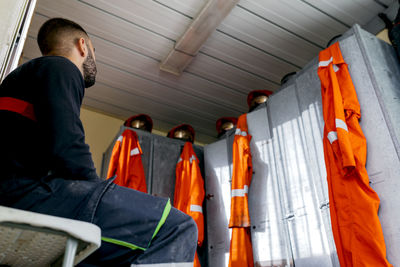 From below unknown firefighter sitting near a row of metal lockers with helmets on top and orange uniform hanging down