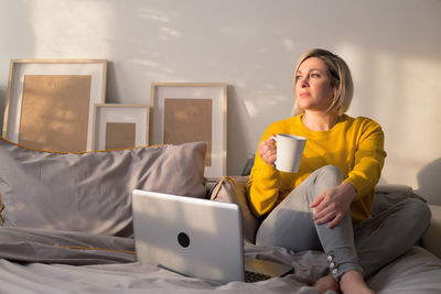 Woman holding coffee cup while sitting on bed at home