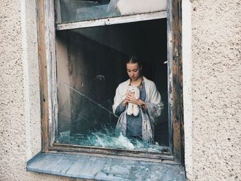 Woman with stuffed toy in building seen through broken window