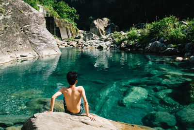 Rear view of shirtless man sitting on rock in front of lake