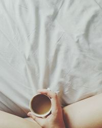 Low section of woman holding coffee cup on bed