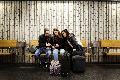 Full length of friends looking at smart phone while sitting against wall on subway station platform