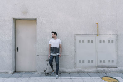 Young male with leg prosthesis smiling away while holding skateboard and standing against concrete building on street