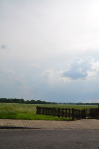 Fence on grassy field in front of road against sky during sunny day