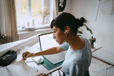 High angle side view of female freelancer with wireless in-ear headphones examining diary at desk while working from hom