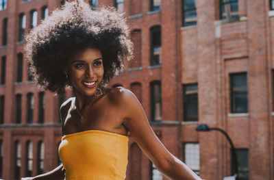 Happy young woman with afro hairstyle standing on bridge against building