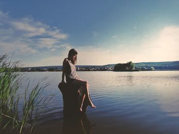 Woman sitting on wooden post over lake during sunset