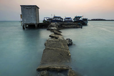 Abandoned pier by boats moored on sea at sunset