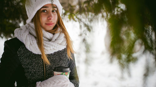 Young woman wearing warm clothing while holding disposable cup amidst trees during winter