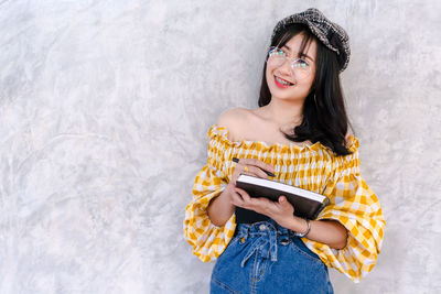 Portrait of smiling young woman using phone while standing against wall