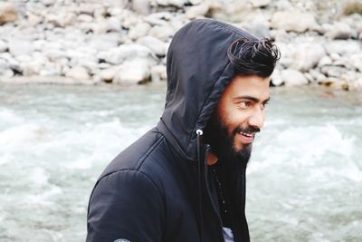 Smiling young man wearing hooded shirt while standing by river