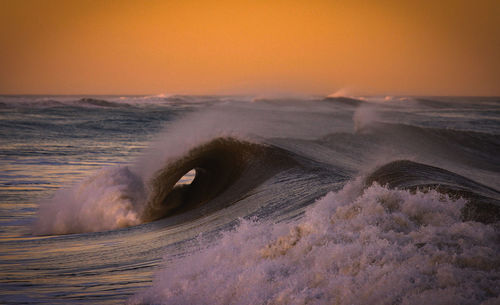 Sea waves splashing on shore against sky during sunset