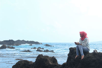 Woman sitting on rock at beach against sky