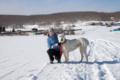 A young girl is sitting next to a big shepherd dog on a snow-white frozen lake against village.