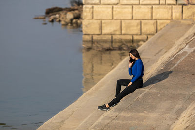 A beautiful teenager talking on her smart phone. she is sitting on the bank of the river. copy space