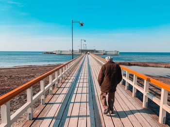 Rear view of mid adult man walking on pier against blue sky