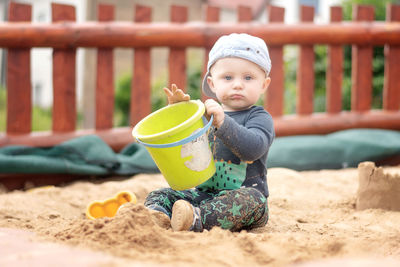 Portrait of cute baby boy holding bucket while sitting on sand in playground