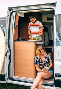 Young man cooking in a camper van while his wife looks at the mobile