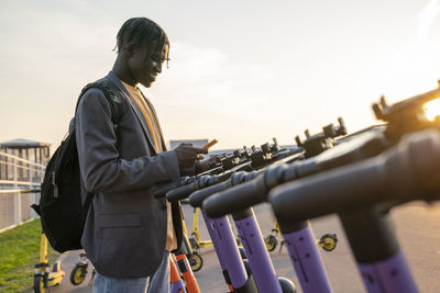 Smiling young businessman using smart phone in front of electric push scooter station