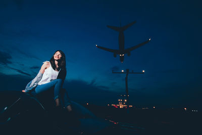 Smiling woman sitting on car with airplane landing at airport against blue sky