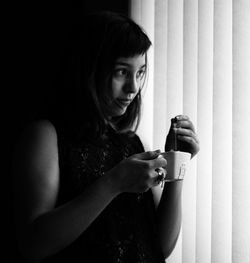 Thoughtful young woman holding tea cup while looking away by window blinds at home