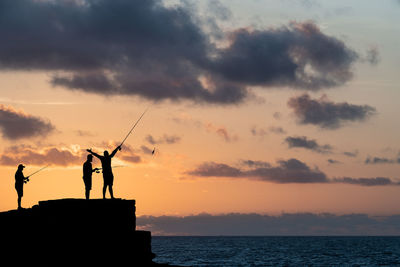 Silhouette people fishing by sea against sky during sunset