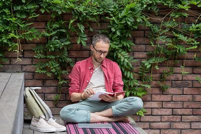 Focused man sits cross-legged outdoors and reads book enjoying novel with exciting plot.