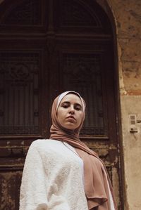 Low angle portrait of young woman wearing hijab standing against door