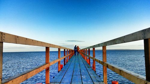 Rear view of couple walking on pier over sea against blue sky