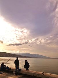 Rear view of silhouette people fishing at sea shore against sunset sky