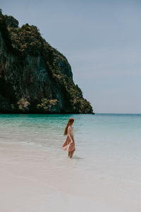 Back view full body of faceless female in dress standing on sandy beach near azure water against rocky cliffs in thailand