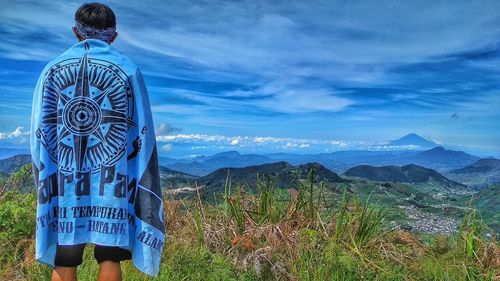 Rear view of man with blue fabric standing on landscape against sky