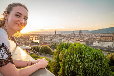 Portrait of young woman in city against sky during sunset