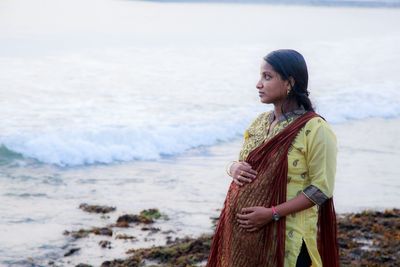 Portrait of young woman standing at beach against sky