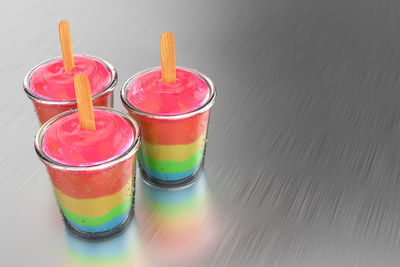 3d rendering of a group of rainbow pudding popsicles with a glass cup on a brush metal background.