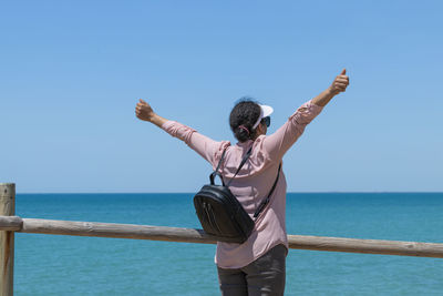Rear view of woman with arms raised against sea against clear blue sky