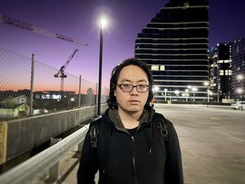 Portrait of young asian man standing on rooftop car park against lights and buildings.