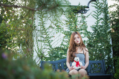 Young woman with face paint holding teddy bear while sitting on bench in yard