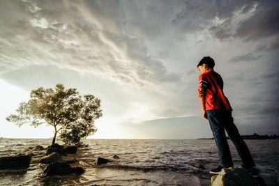 Boy standing on rock in sea against cloudy sky during sunset