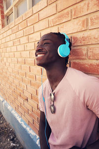 Smiling teenage boy listening music while standing against brick wall