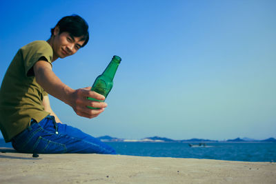Man holding beer bottle sitting on retaining wall against blue sky