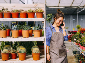 Happy lady in apron smiling and answering phone call while leaning on shelves with succulents during work in hothouse