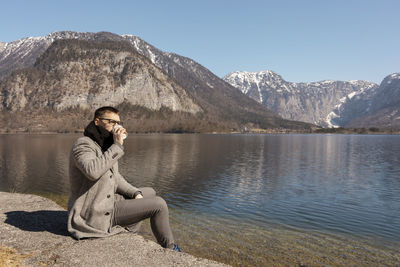 Young adult man sitting outdoors, drinking coffee and enjoying mountains, lake, good weather