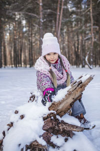Happy girl in knitted hat enjoys winter season and sits in the snow in forest on sunny winter day