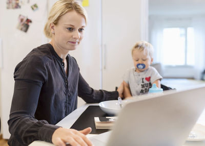 Woman working on laptop while taking care of baby boy at dining table