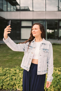 Young woman in white denim jacket taking selfie on street. office building in the background