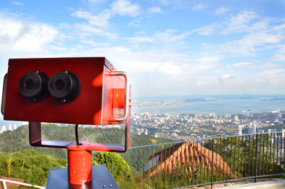 Close-up of red coin-operated binoculars over city against sky